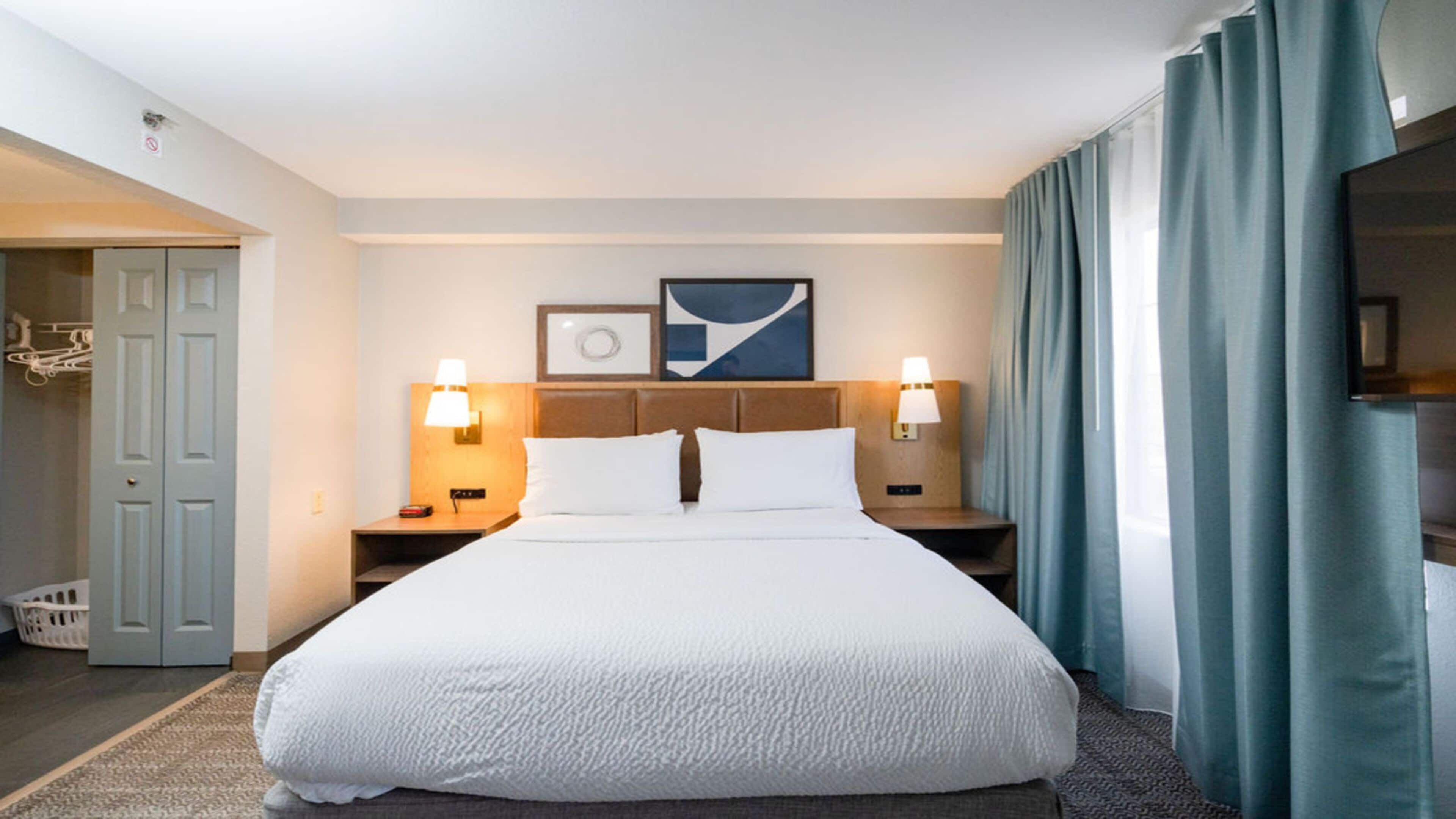 A hotel room with a large bed, two bedside tables with lamps, a headboard with a picture, and a view of the outside through a window with curtains.