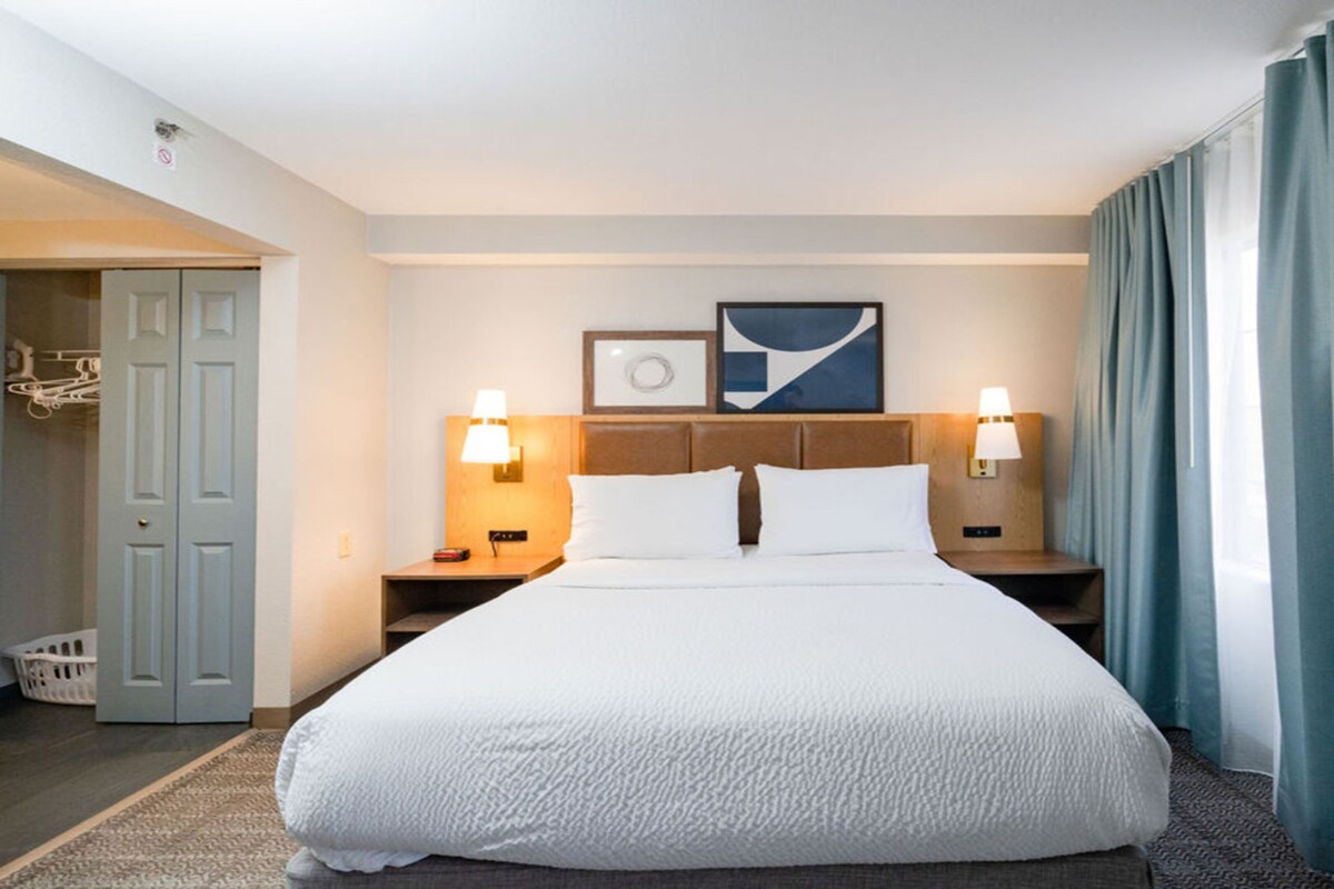 A hotel room with a large bed, two bedside tables with lamps, a headboard with a picture, and a view of the outside through a window with curtains.