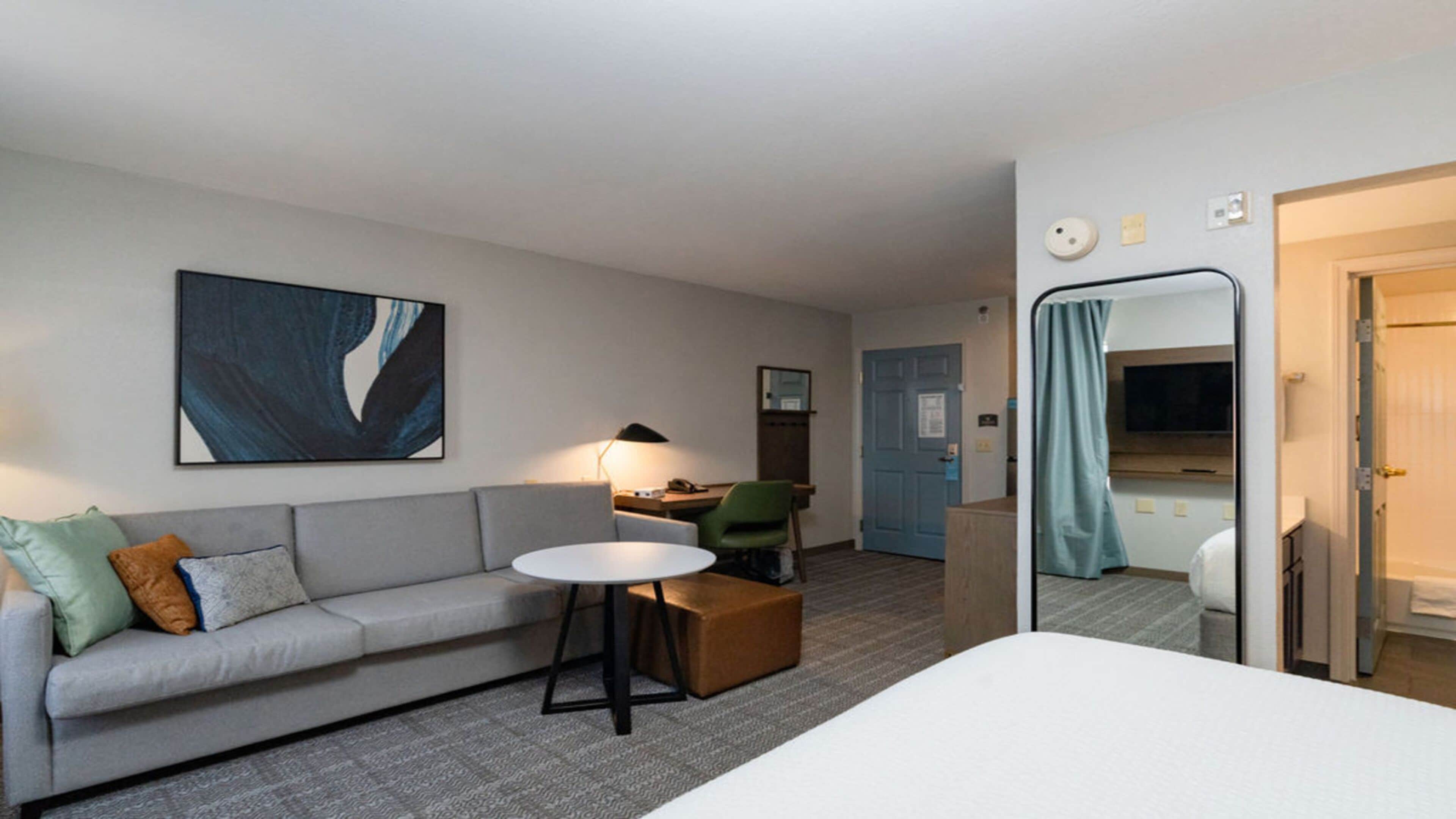 A hotel room with a grey sofa, a small round table, a green chair, a large mirror, and a bed.