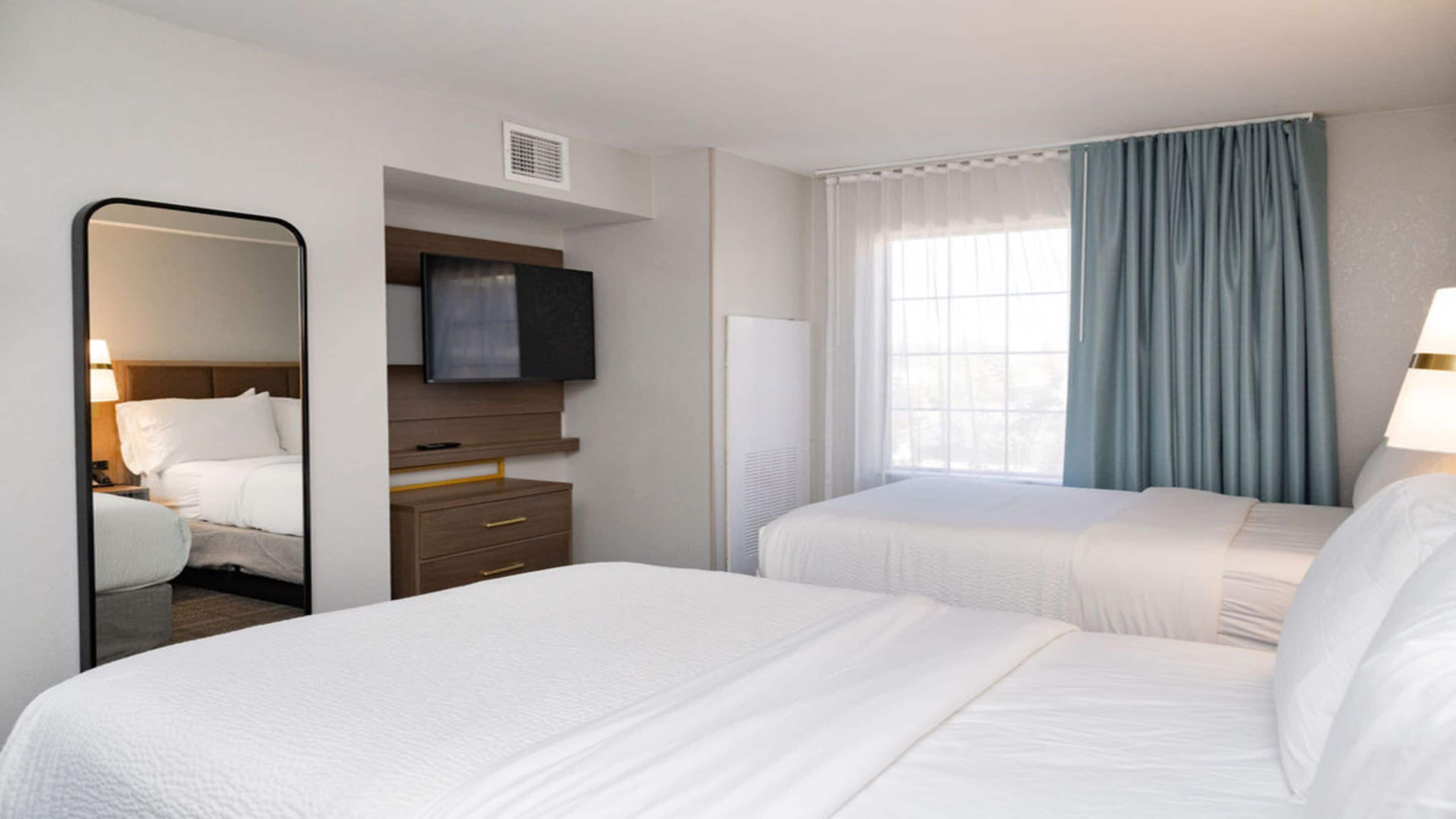A hotel room with two beds, a flat-screen TV, a mirror, a dresser, and a window with curtains.