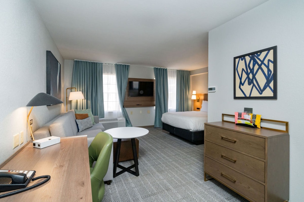 A hotel room with a bed, a desk with a lamp, a chair, a dresser, and a wall-mounted TV.