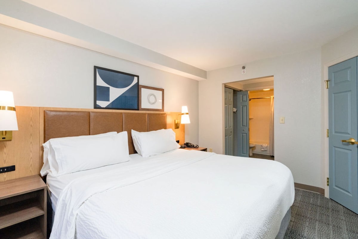 A hotel room with a large bed, two bedside lamps, a wooden headboard, and a framed picture on the wall.