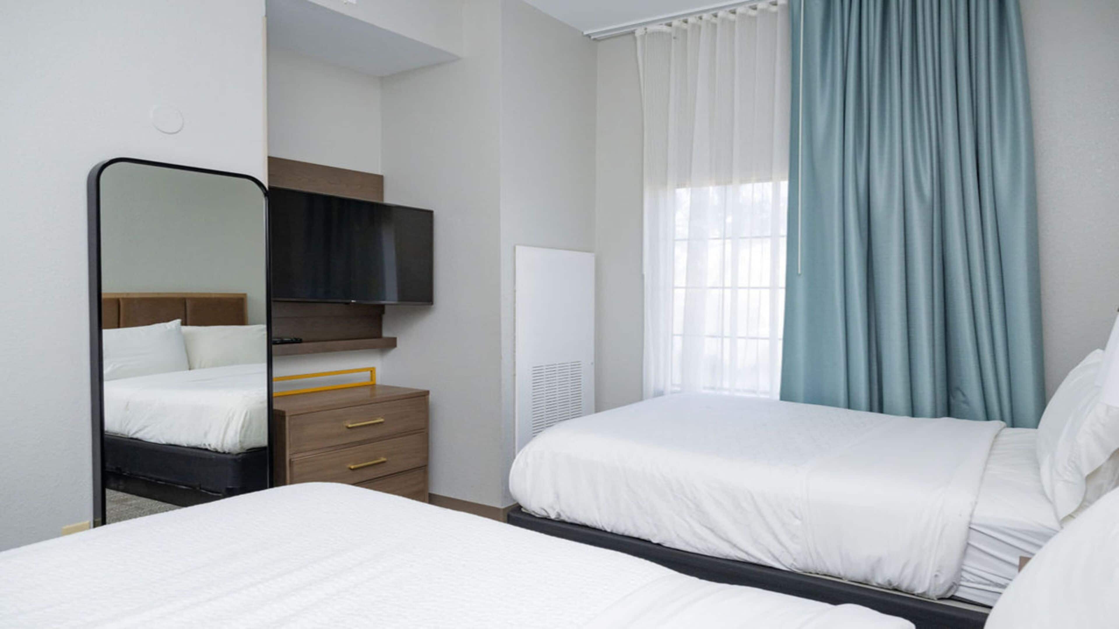 A hotel room with two beds, a flat-screen TV, a wooden dresser, and a window with curtains.