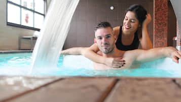 A couple in a hot tub with a waterfall feature, a tiled wall, and a window in the background.