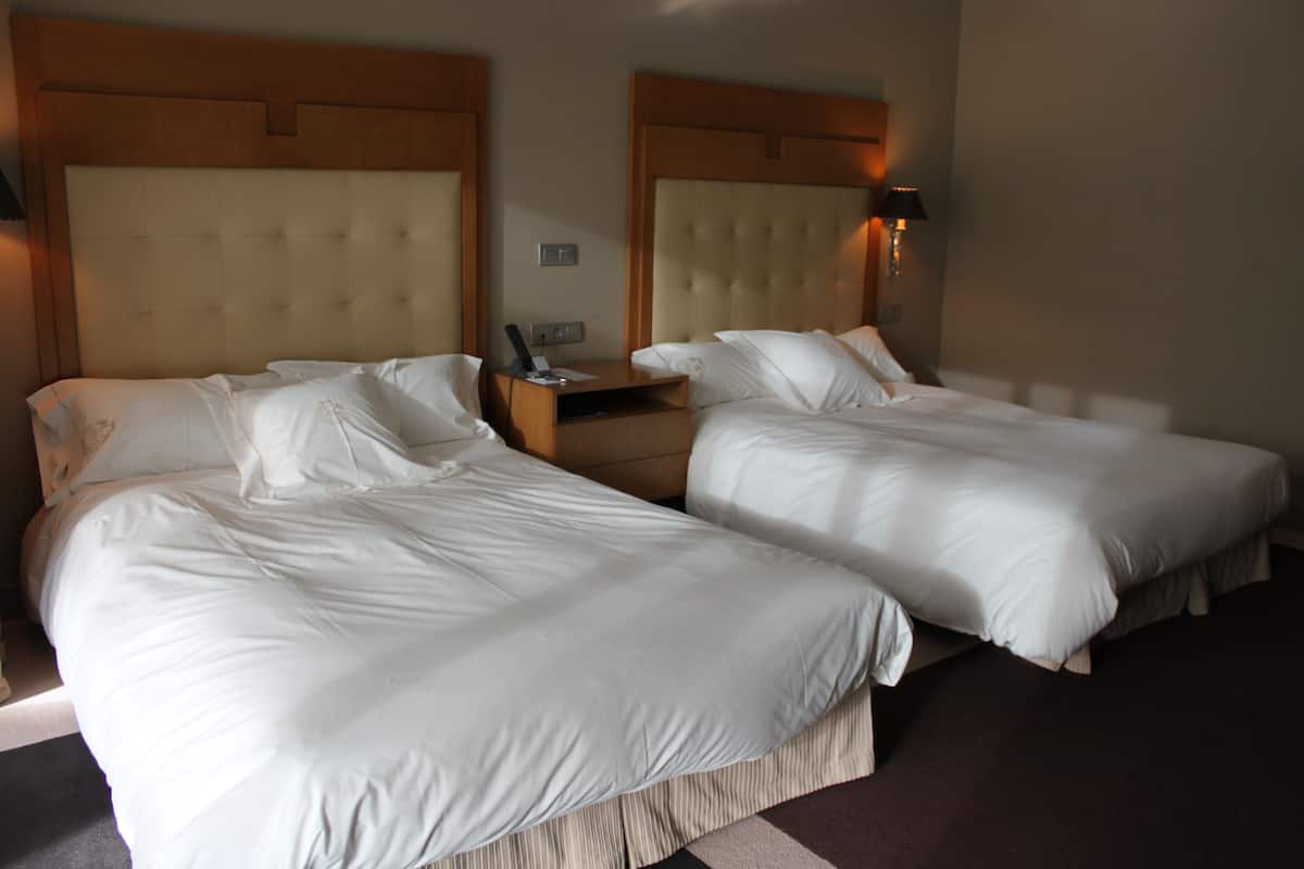 A hotel room with two single beds, a wooden headboard, a bedside table with a lamp, and a small clock.