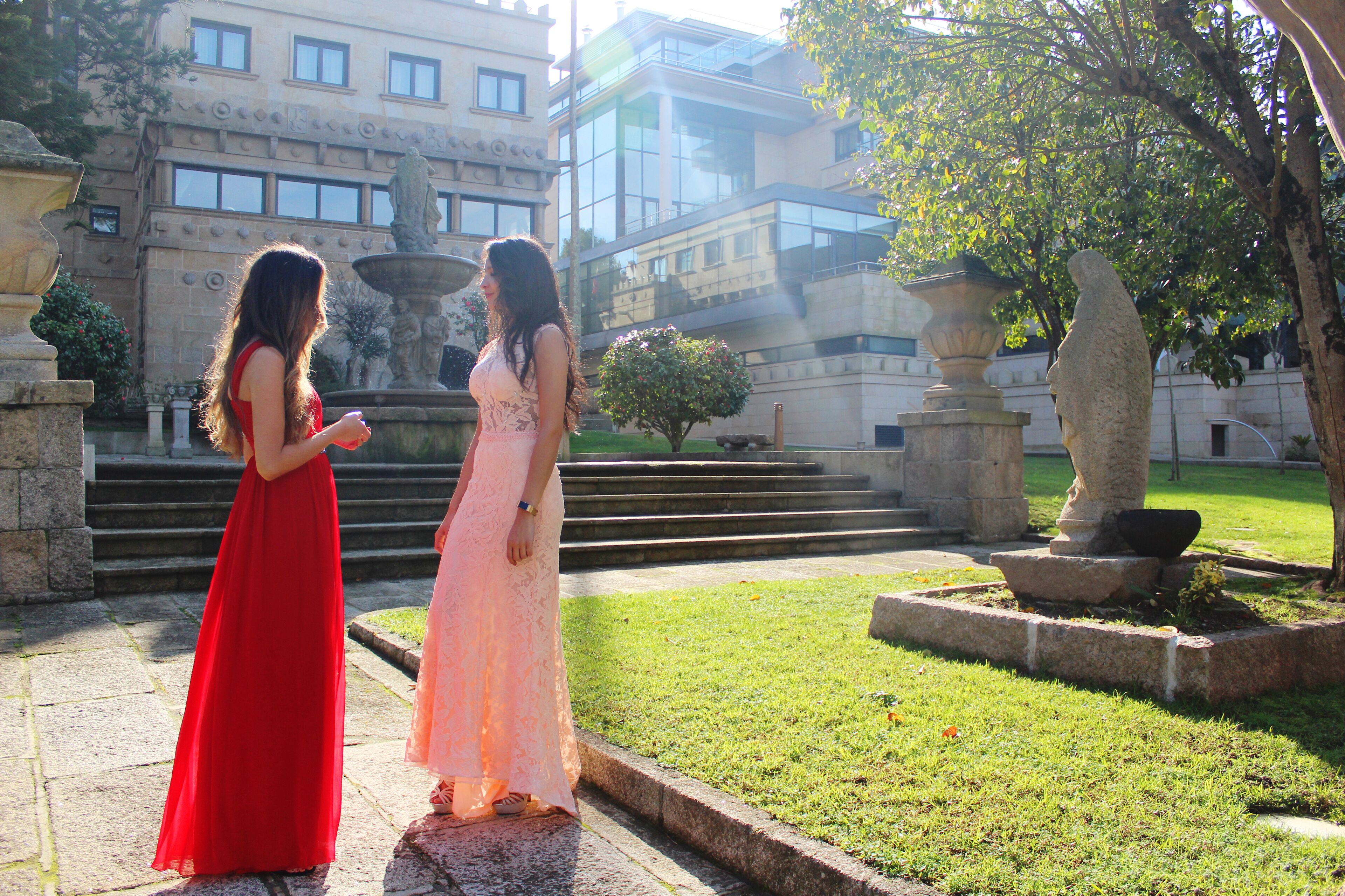 Dos mujeres con vestidos largos de pie en un patio con una fuente, escalones y un edificio al fondo.