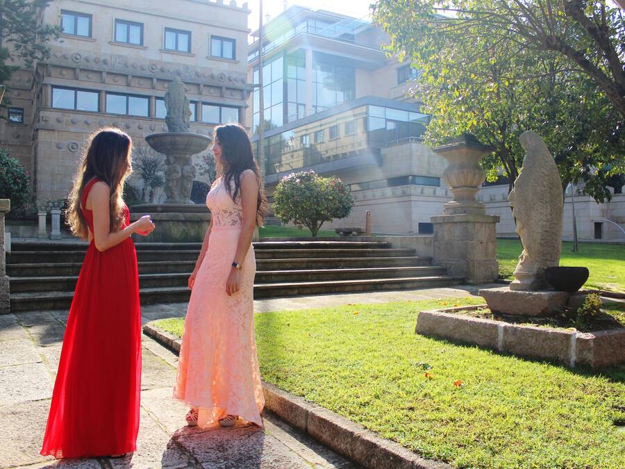 Dos mujeres con vestidos largos de pie en un patio con una fuente, escalones y un edificio al fondo.