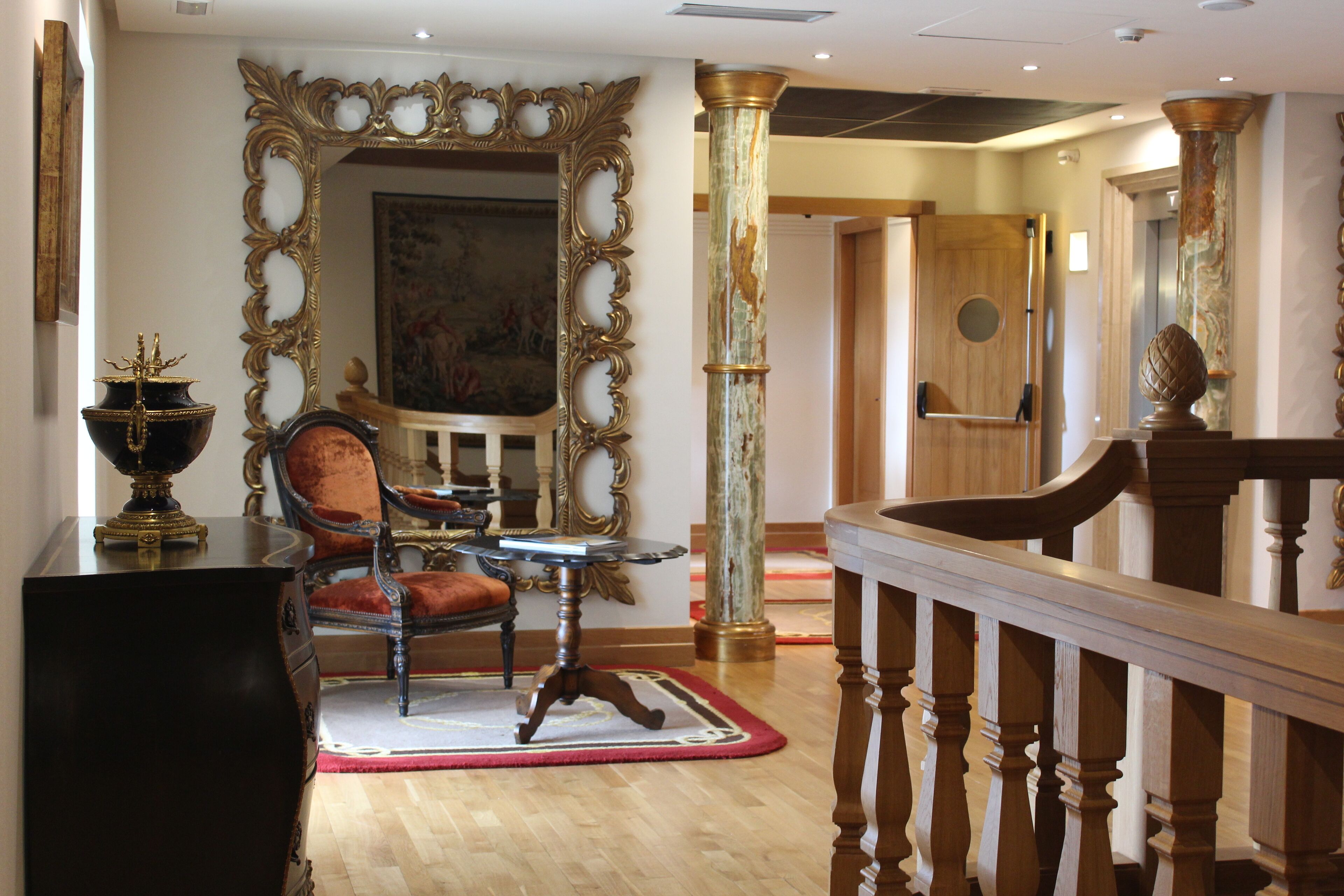 Una estancia amplia con un espejo grande y ornamentado, un sillón tapizado en rojo, una mesa de madera y una escalera con barandilla.