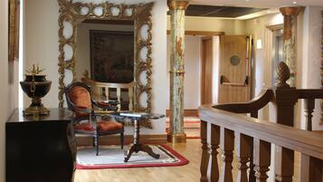 Una estancia amplia con un espejo grande y ornamentado, un sillón tapizado en rojo, una mesa de madera y una escalera con barandilla.