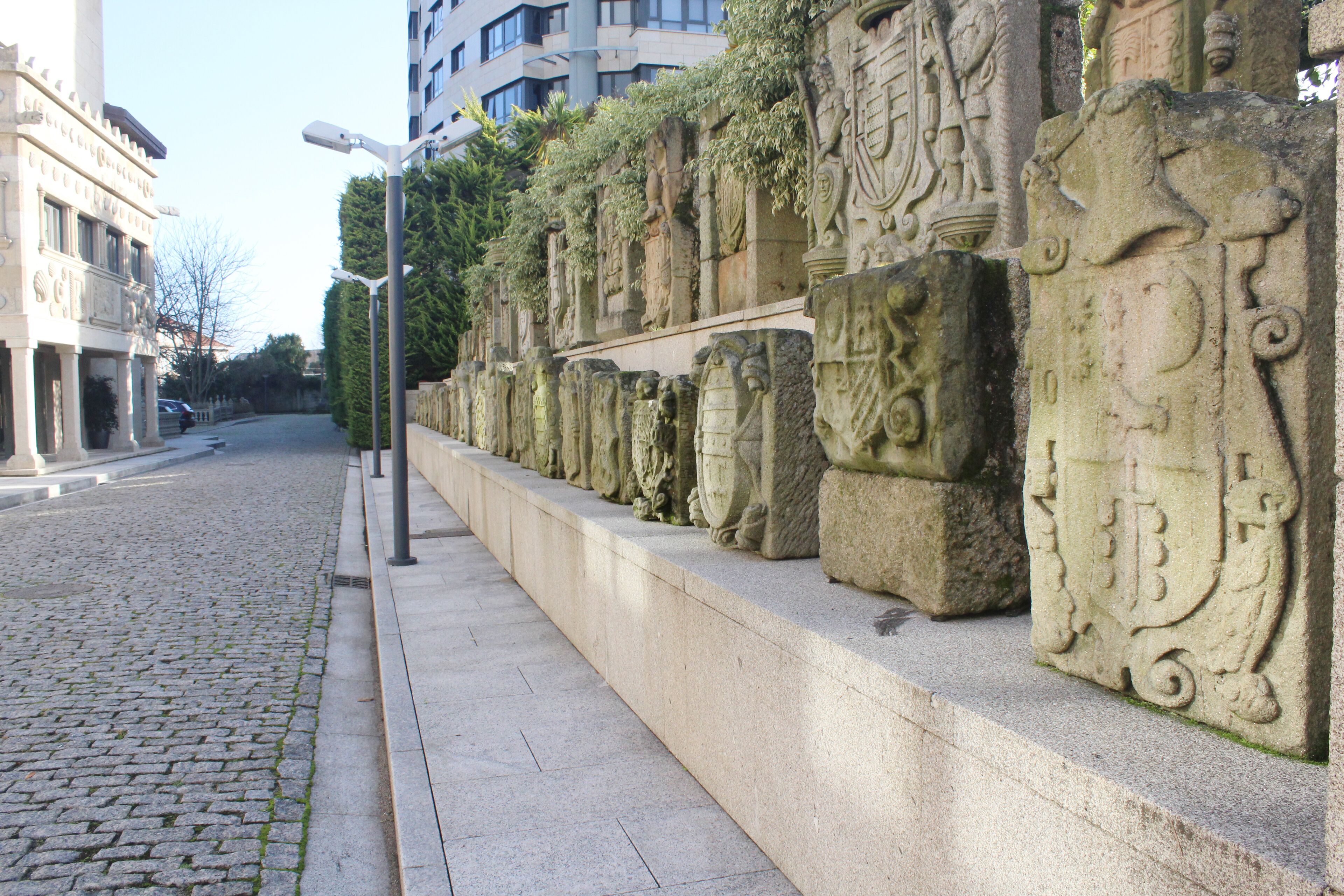 Une rue pavée bordée d’une rangée de sculptures anciennes en pierre et d’un lampadaire moderne.