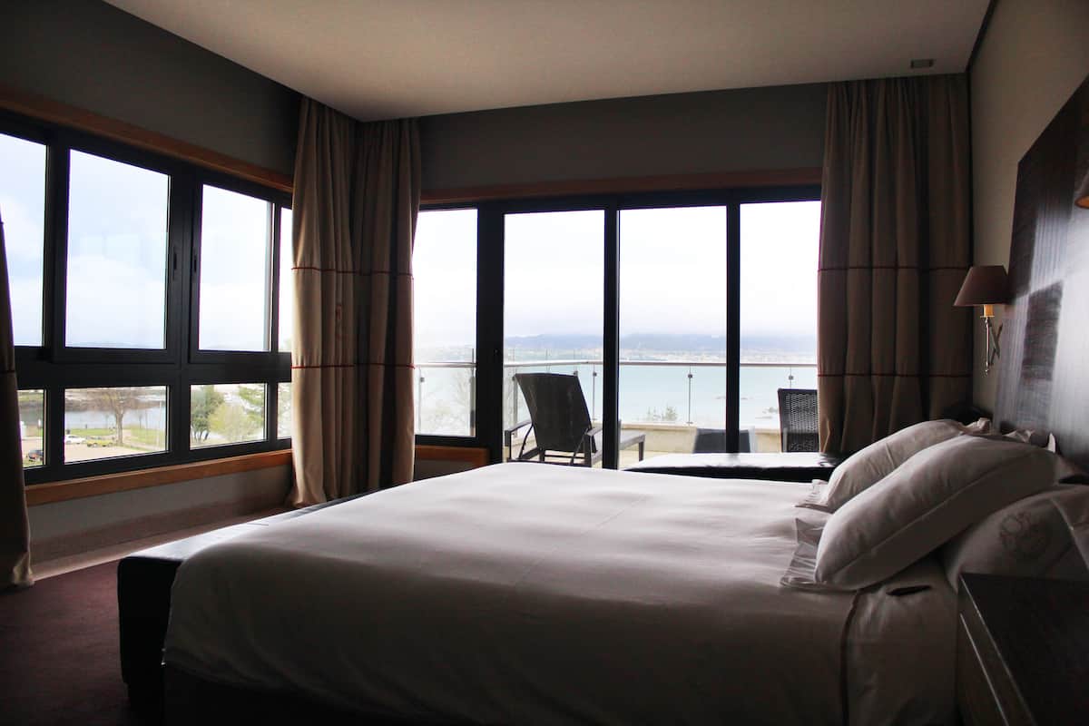 A hotel room with a large bed, a chair, a table, and a view of the city and mountains through large windows.