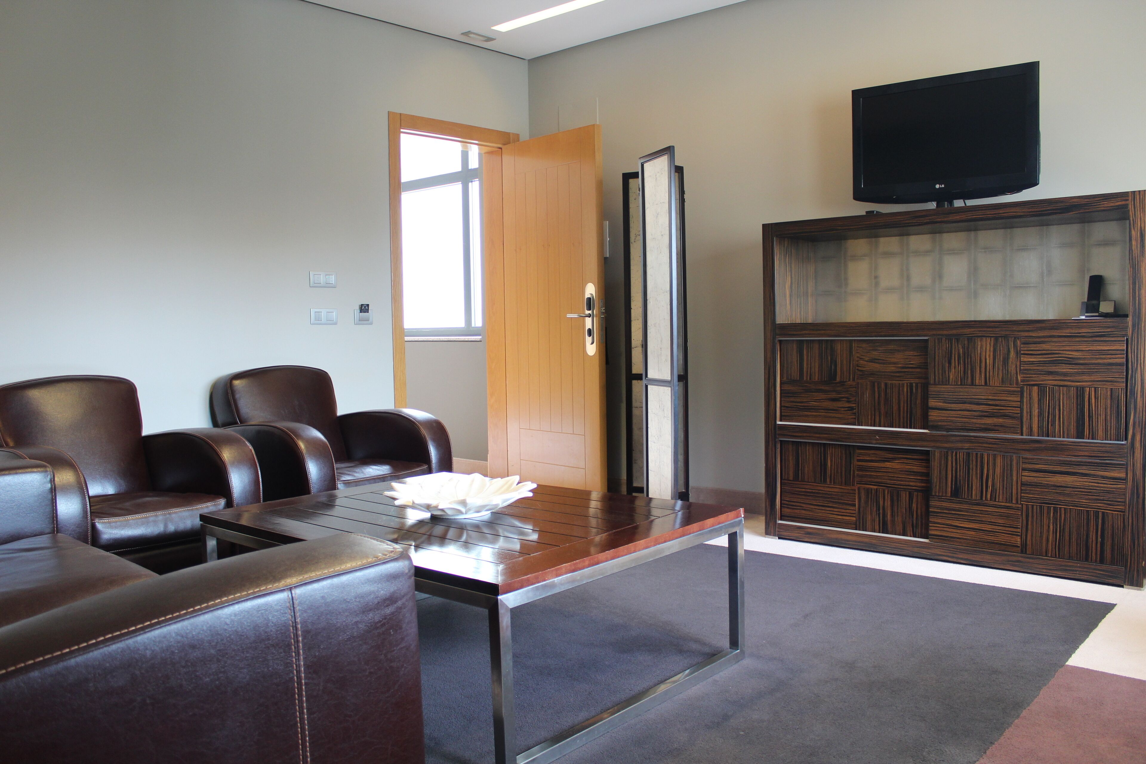 Un salón de conferencias con sillas de cuero, una mesa con superficie de vidrio, un mueble de madera con un televisor y una puerta con ventana.