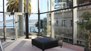 A modern interior with large windows, a black ottoman, and a view of a coastal area.