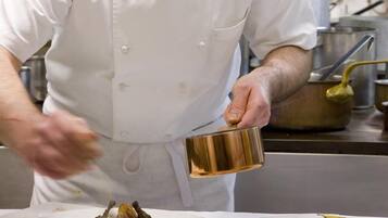 Un chef, vêtu d’un uniforme blanc, prépare des plats dans une cuisine équipée de luminaires suspendus blancs et d’appareils électroménagers en acier inoxydable.