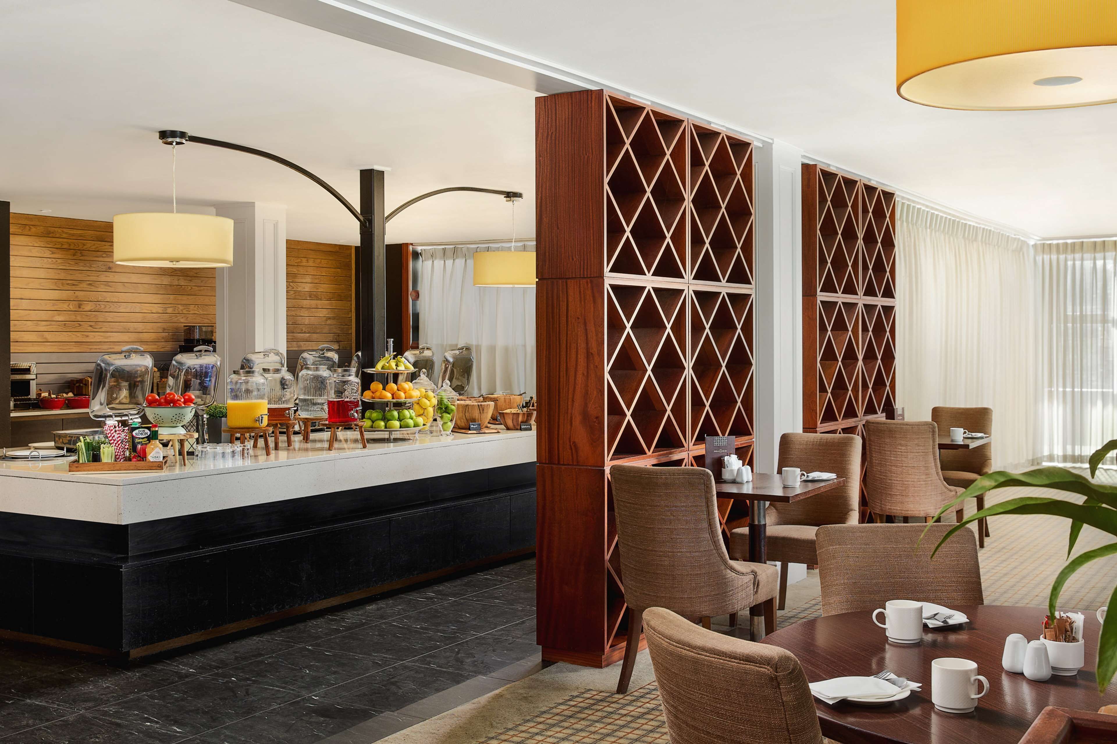 Ein Hotel-Frühstücksbereich mit einer Theke, die Früchte und Getränke präsentiert, Sitzmöglichkeiten und einem dekorativen Holztrennwerk.