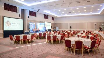 Ett stort konferensrum med flera runda bord och röda stolar, en stor skärm som visar en presentation och en scen.