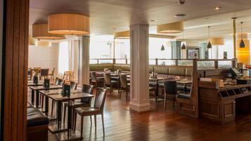 Un restaurante con mesas y sillas de madera, una zona de bar y grandes ventanales que dejan entrar luz natural.