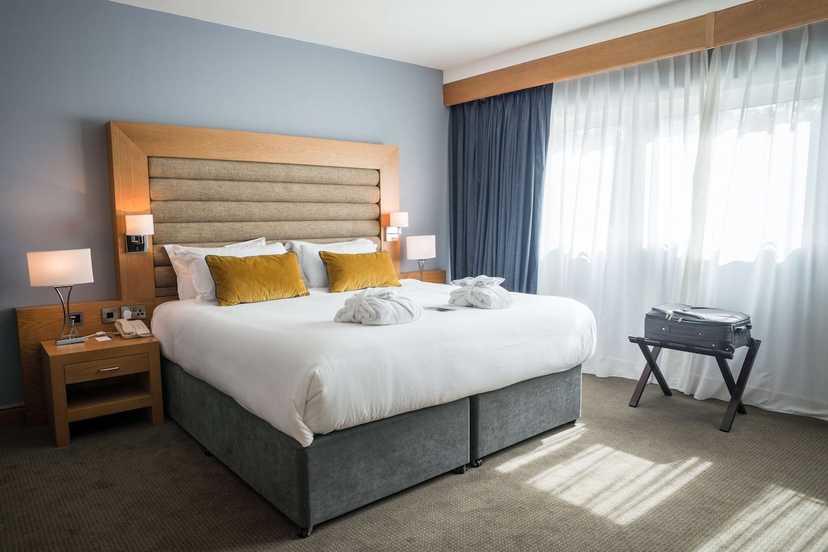 Habitación de hotel con una cama grande, dos mesitas de noche con lámparas, una maleta sobre un atril y una ventana con cortinas.