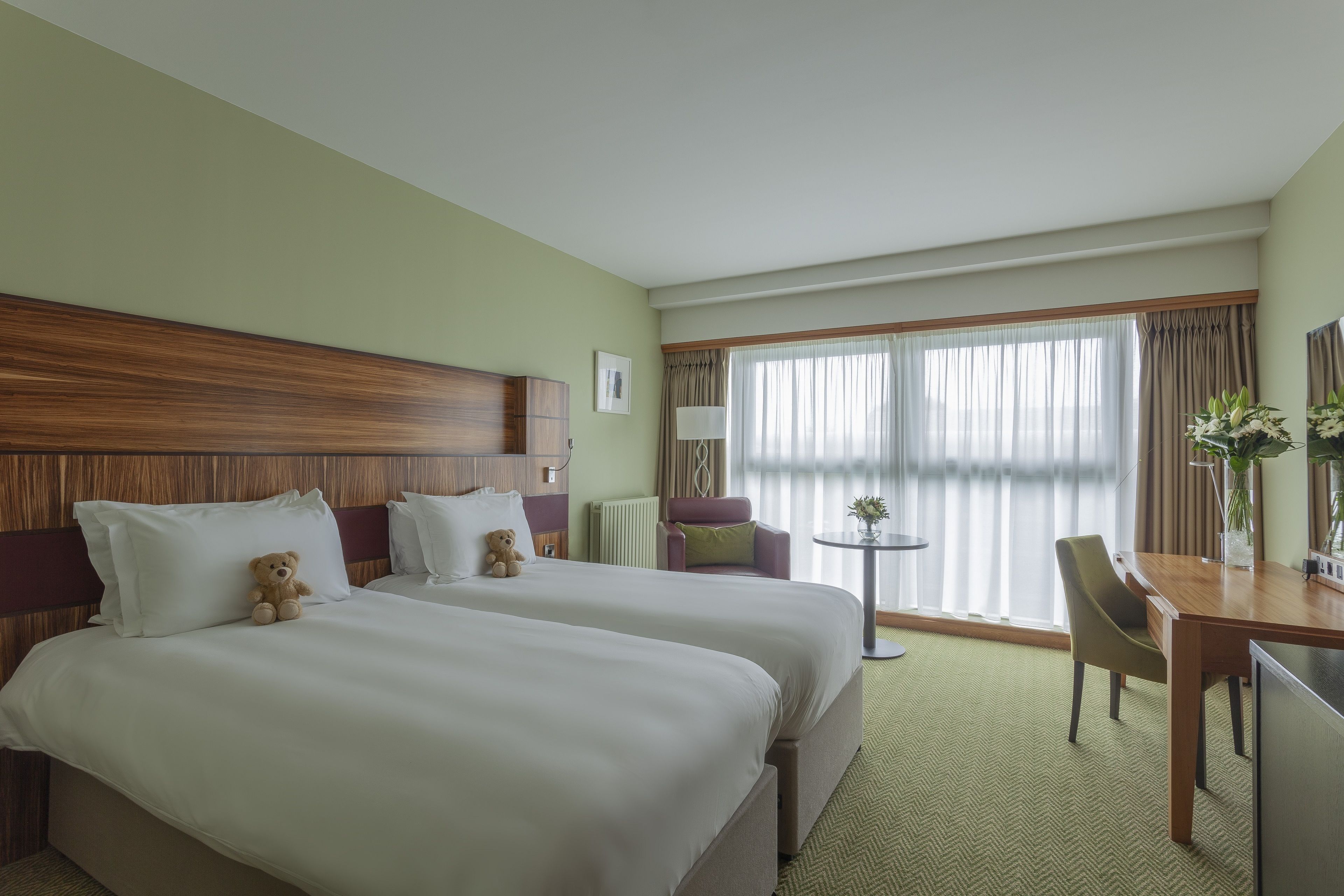 A hotel room with two beds, a dining table, a chair, a small table with a lamp, and a window with curtains.