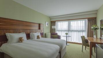 A hotel room with two beds, a dining table, a chair, a small table with a lamp, and a window with curtains.
