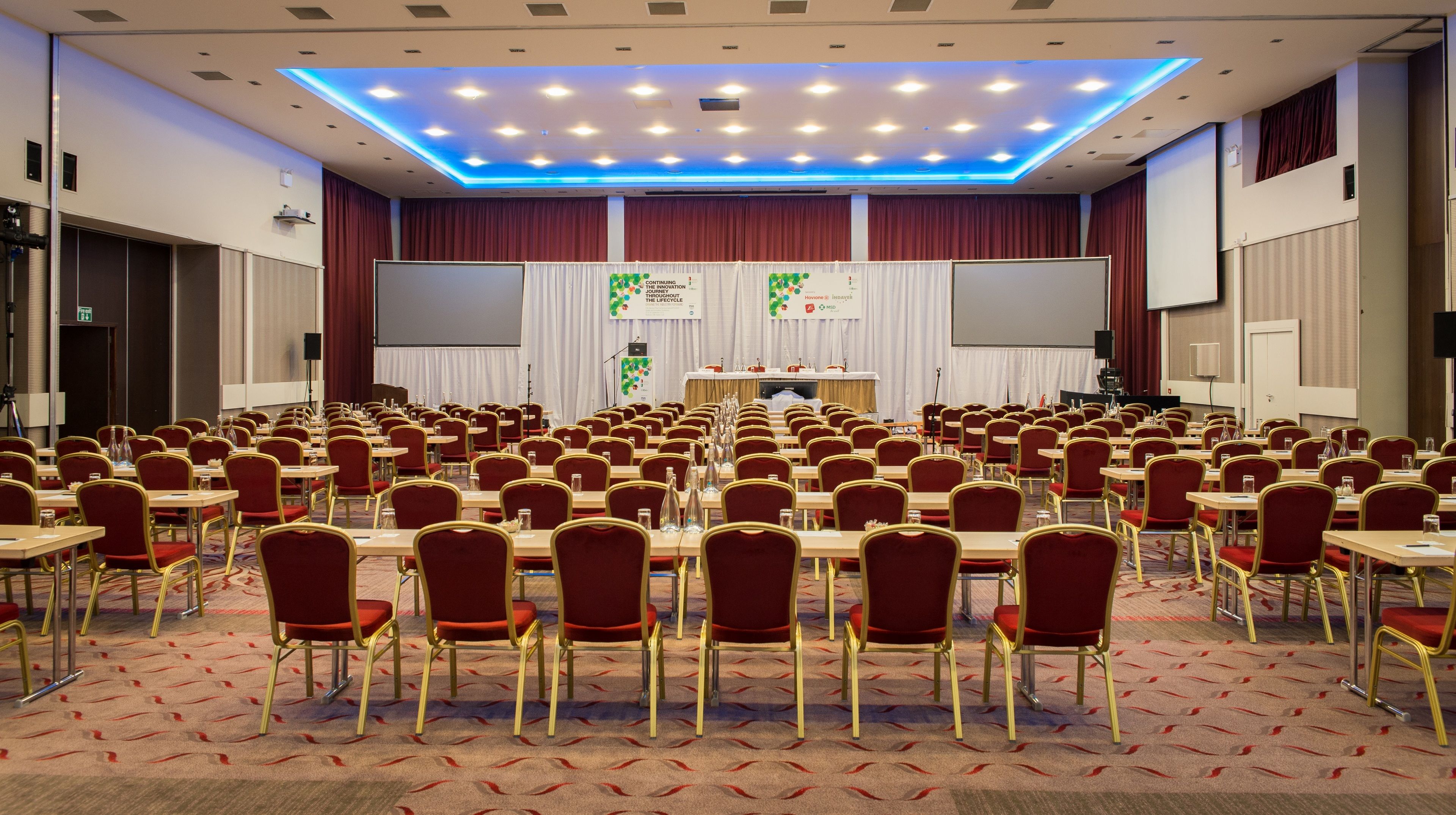 A conference room with rows of tables and chairs, a stage with a podium, and a large screen.
