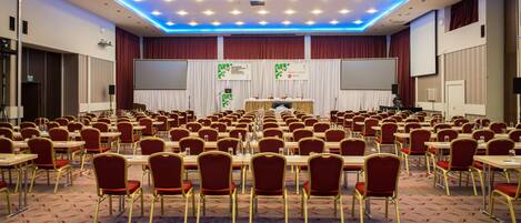 A conference room with rows of tables and chairs, a stage with a podium, and a large screen.