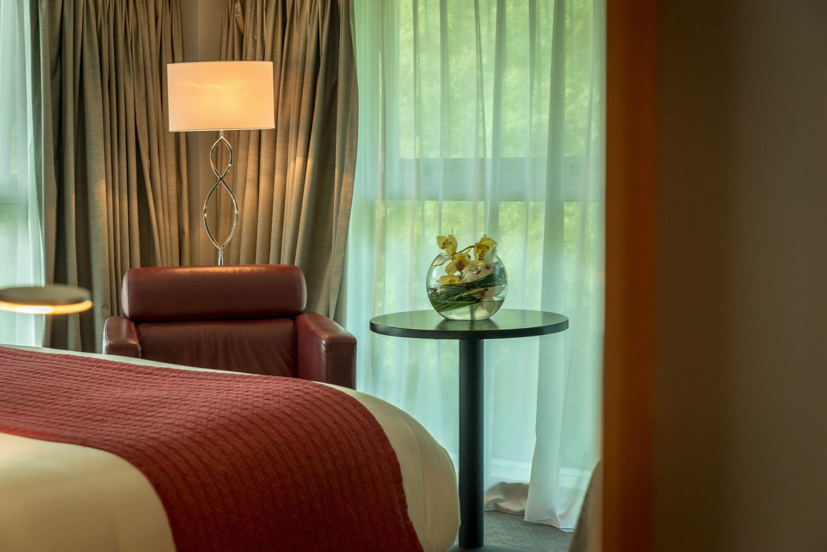 Habitación de hotel con cama, una silla, una lámpara, una mesa con flores y vista a follaje a través de cortinas traslúcidas.