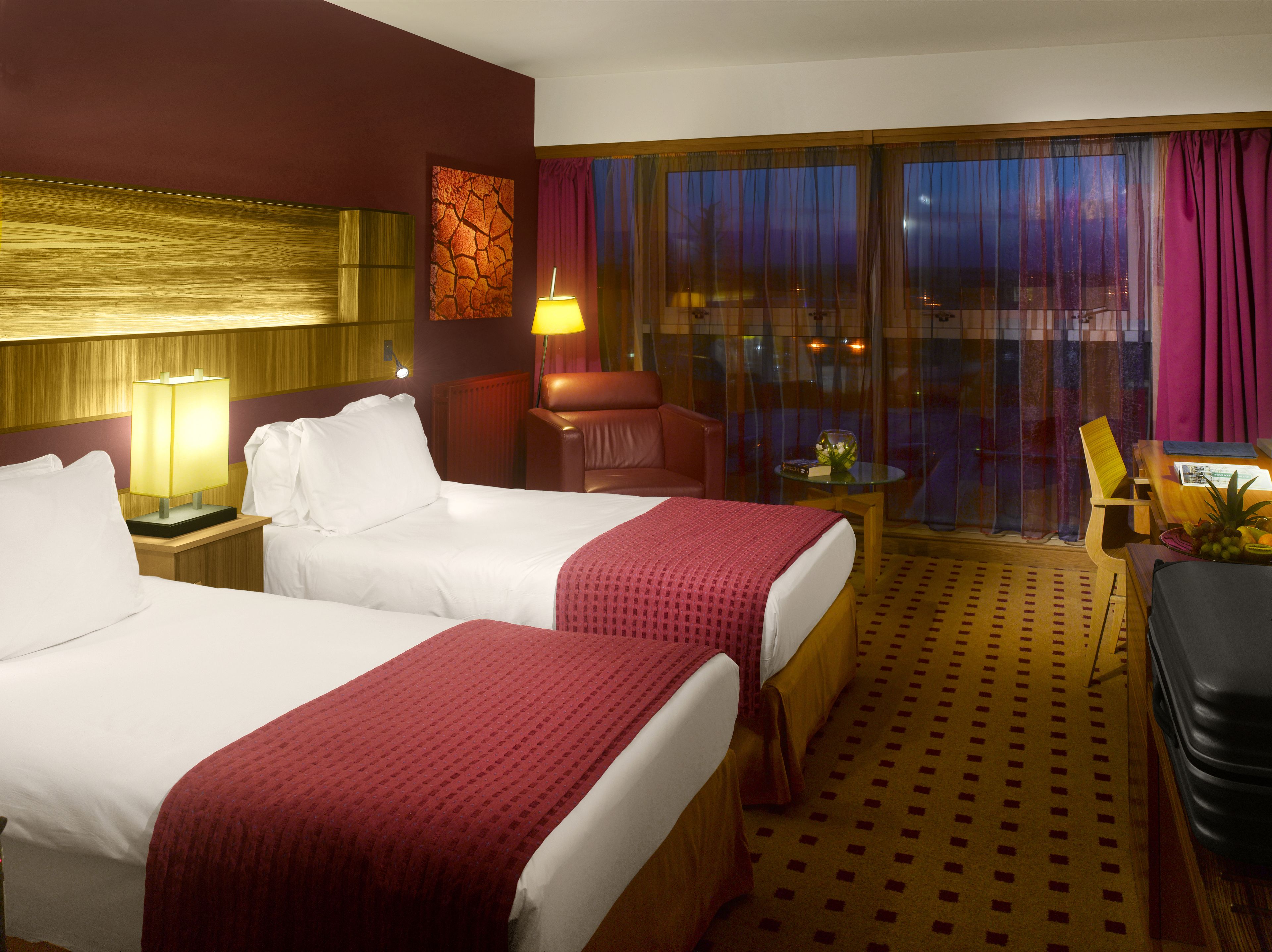 A hotel room with two beds, a desk, a chair, a lamp, and a view of the city through large windows.