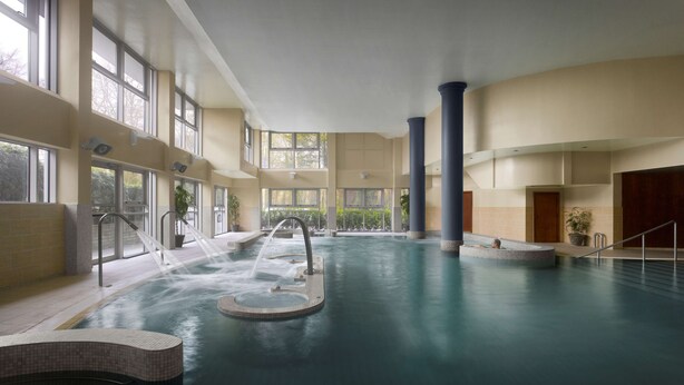 Una piscina interior con varios puntos de acceso, grandes ventanales y una fuente central de agua.