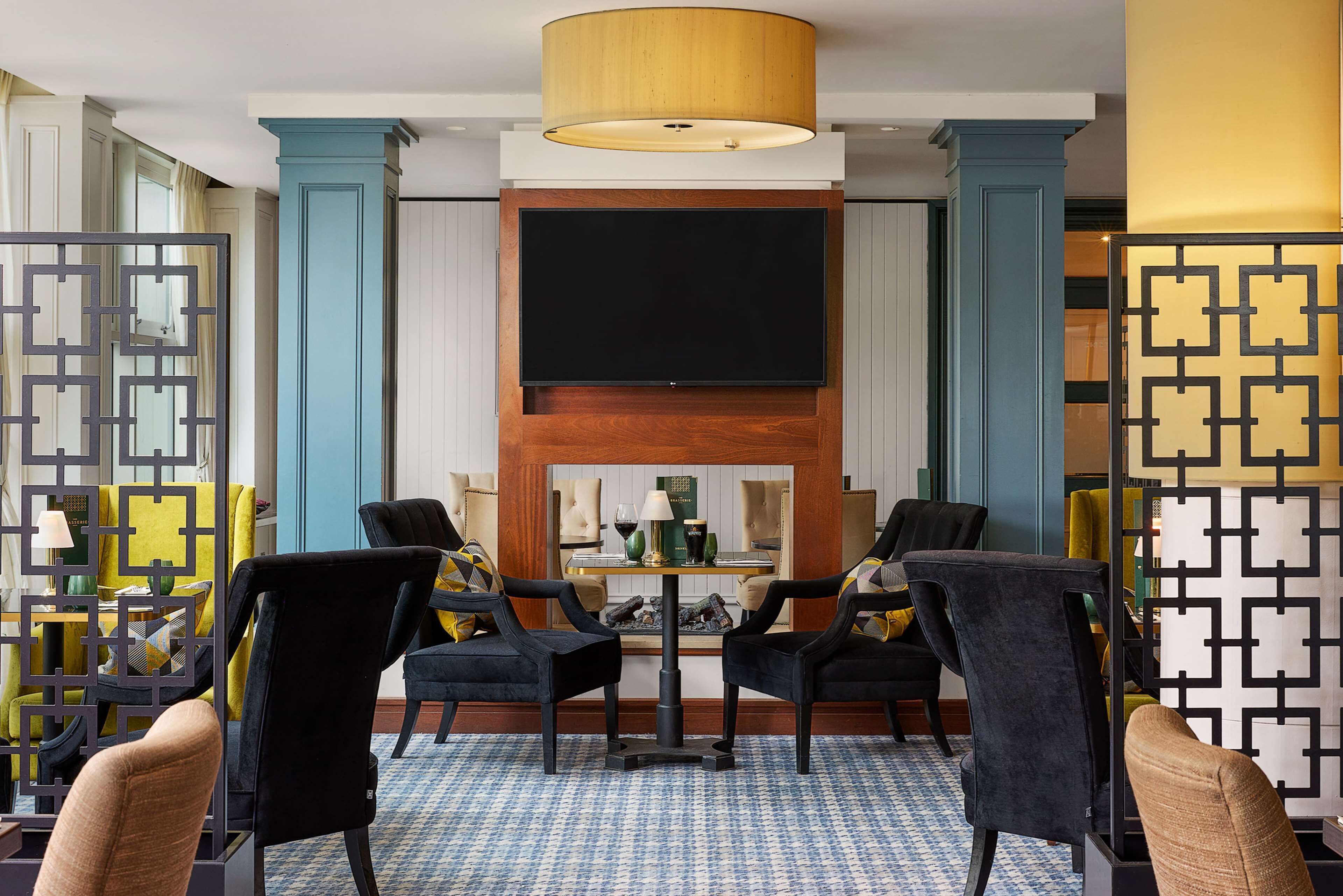 A hotel lobby with a large TV screen, two black chairs, a small table, and a patterned floor.
