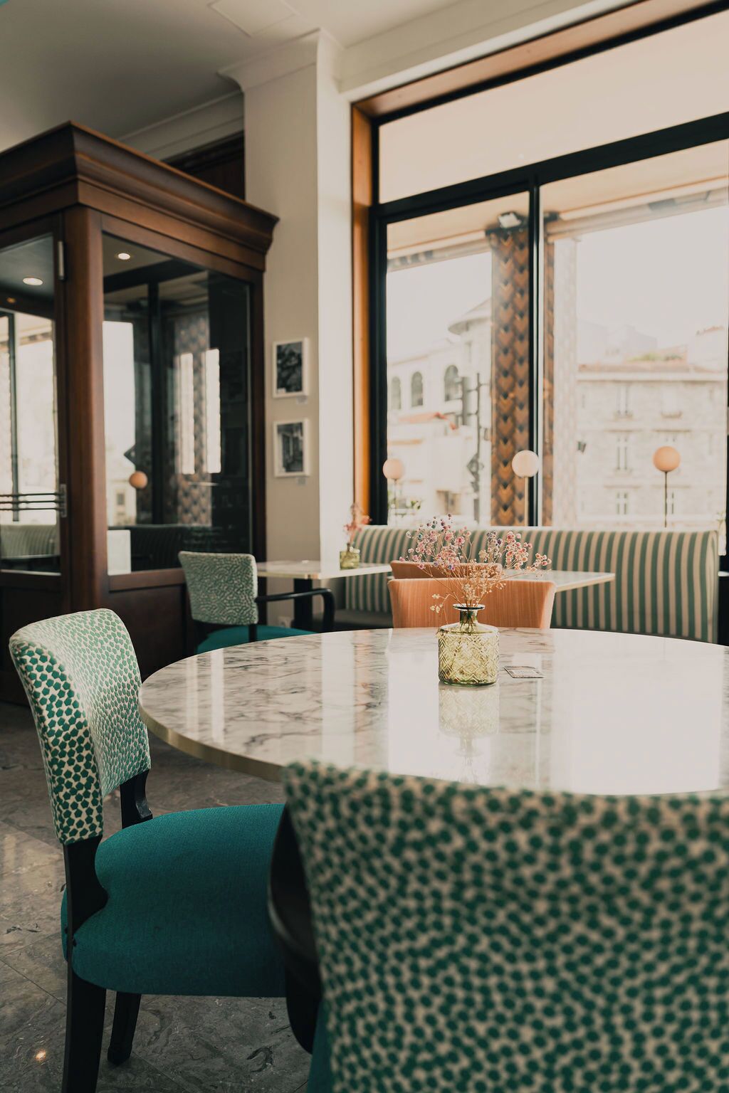 Een ronde tafel met stoelen, een vaas met bloemen en uitzicht op gebouwen door grote ramen.