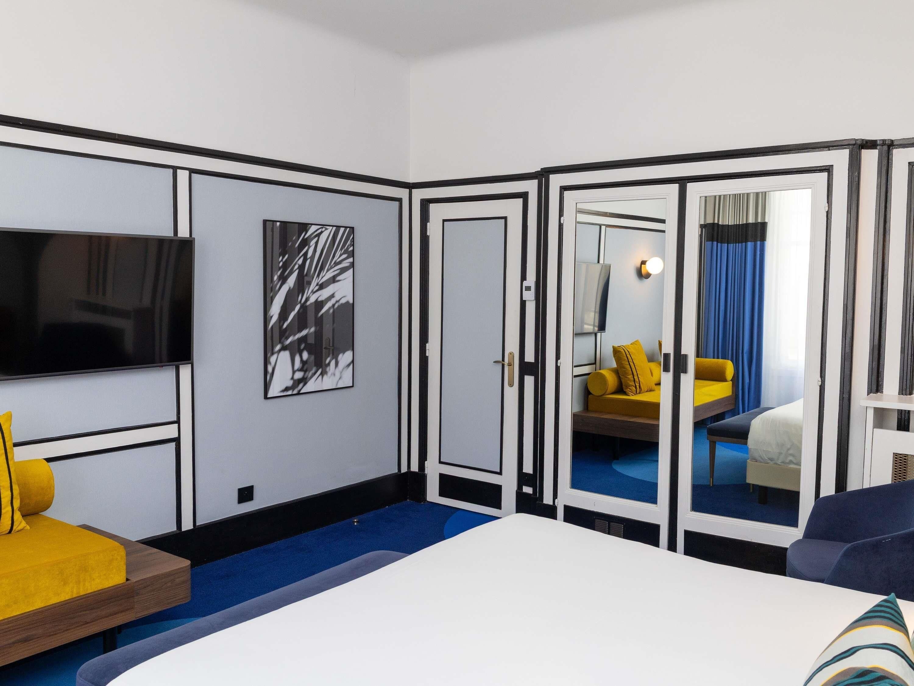 A hotel room with a bed, a TV, a mirror, and a yellow sofa.