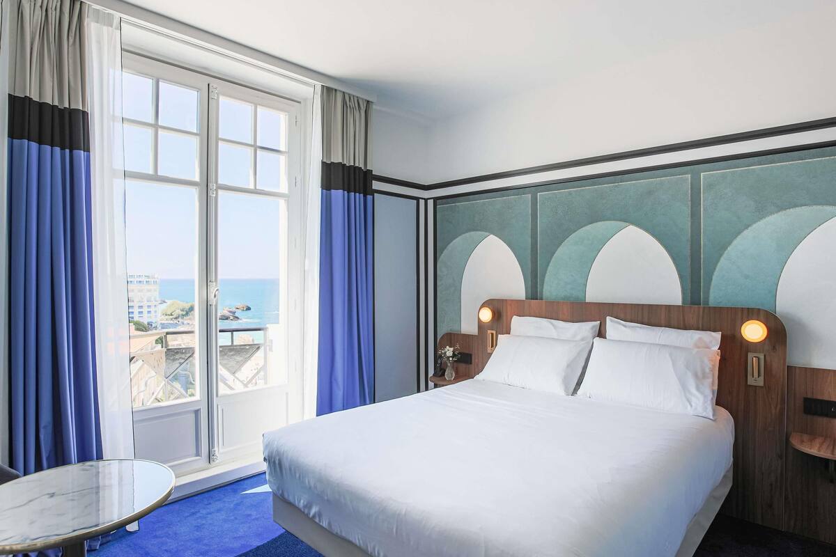 A hotel room with a large bed, a small table, and a view of the sea.