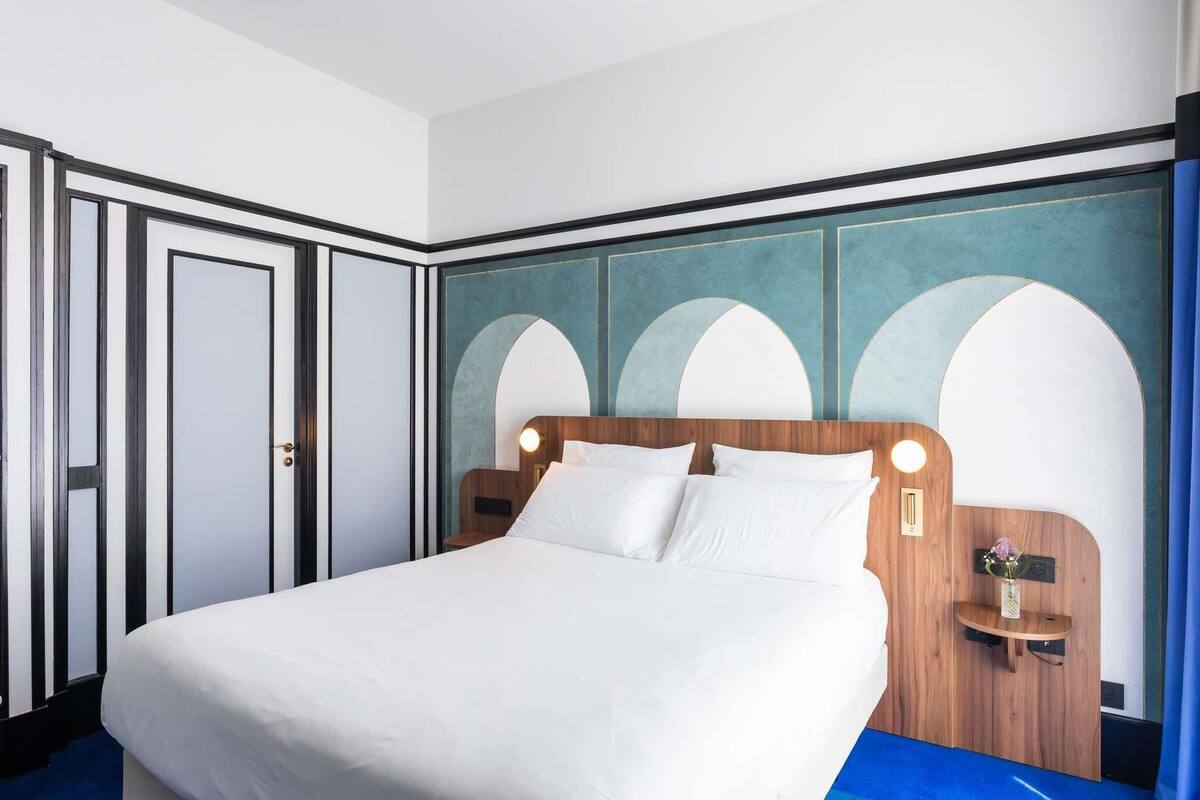 A hotel room with a large bed, a wooden headboard, a nightstand, and a wall with arched designs.