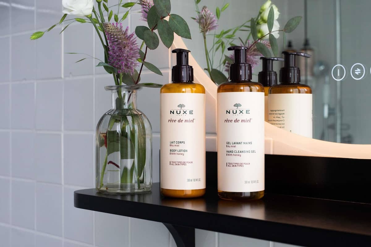 A bathroom with a mirror reflecting three Nuxe body care products, a glass vase with flowers, and a black countertop.