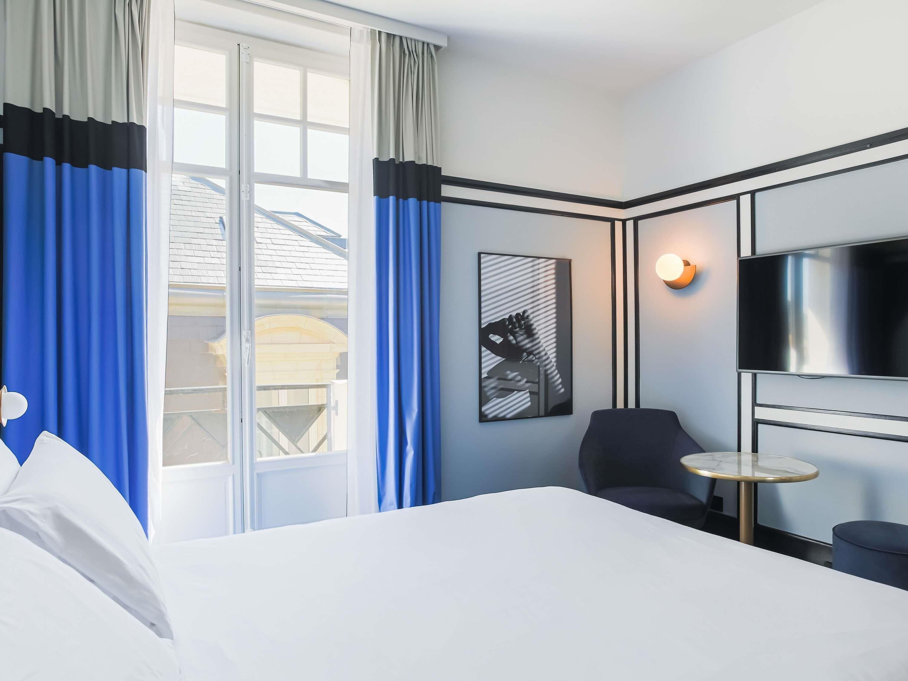 A hotel room with a bed, a TV, a chair, a small table, and a window with curtains.
