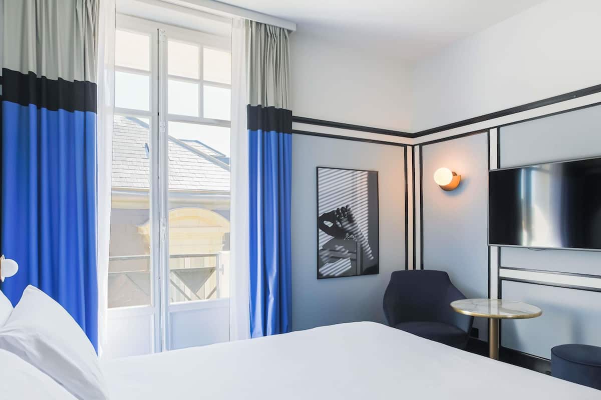 A hotel room with a bed, a TV, a chair, a small table, and a window with curtains.