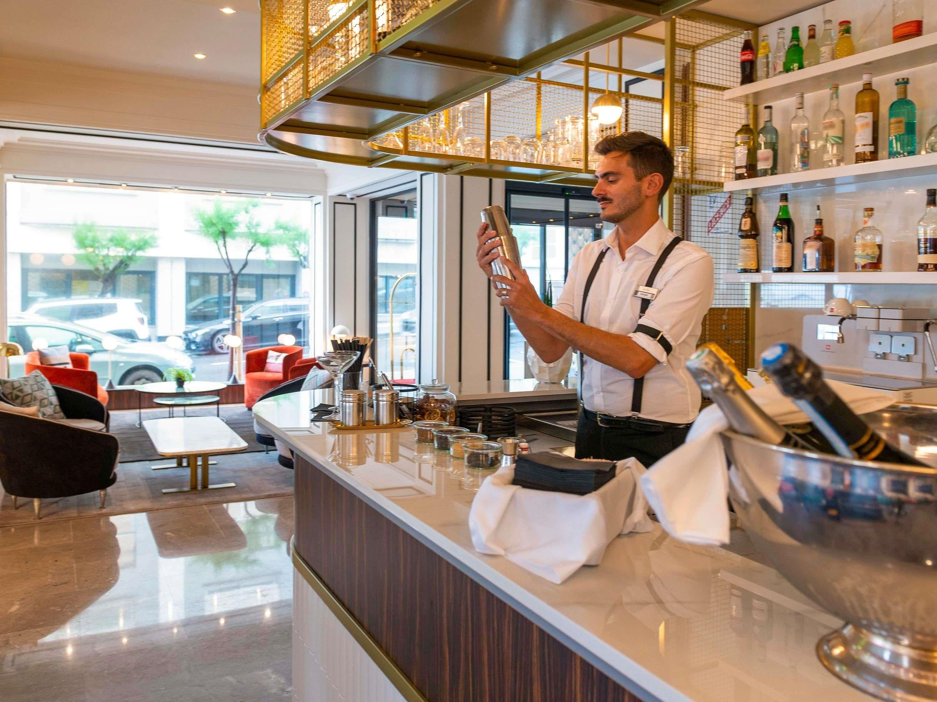Un espace bar avec un barman, un comptoir où sont exposées différentes bouteilles, et un coin salon avec une chaise rouge et une table.