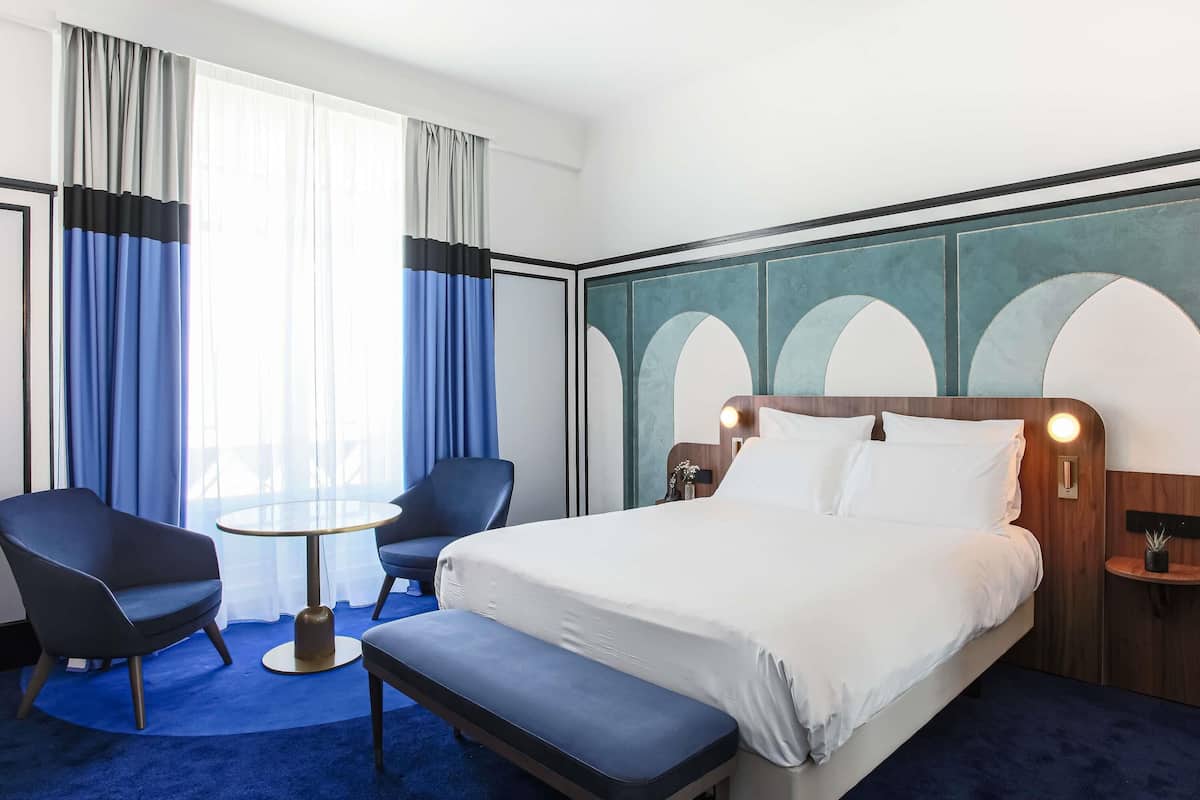 A hotel room with a bed, two chairs, a small table, and a bench.