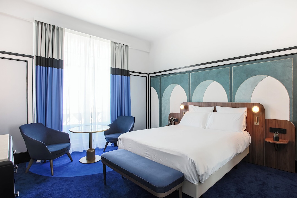 A hotel room with a large bed, a desk with a chair, a small table, and a bench.