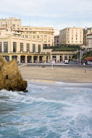 A coastal hotel with a beachfront view, featuring a large rock formation in the foreground, a sandy beach, and a promenade with people walking.