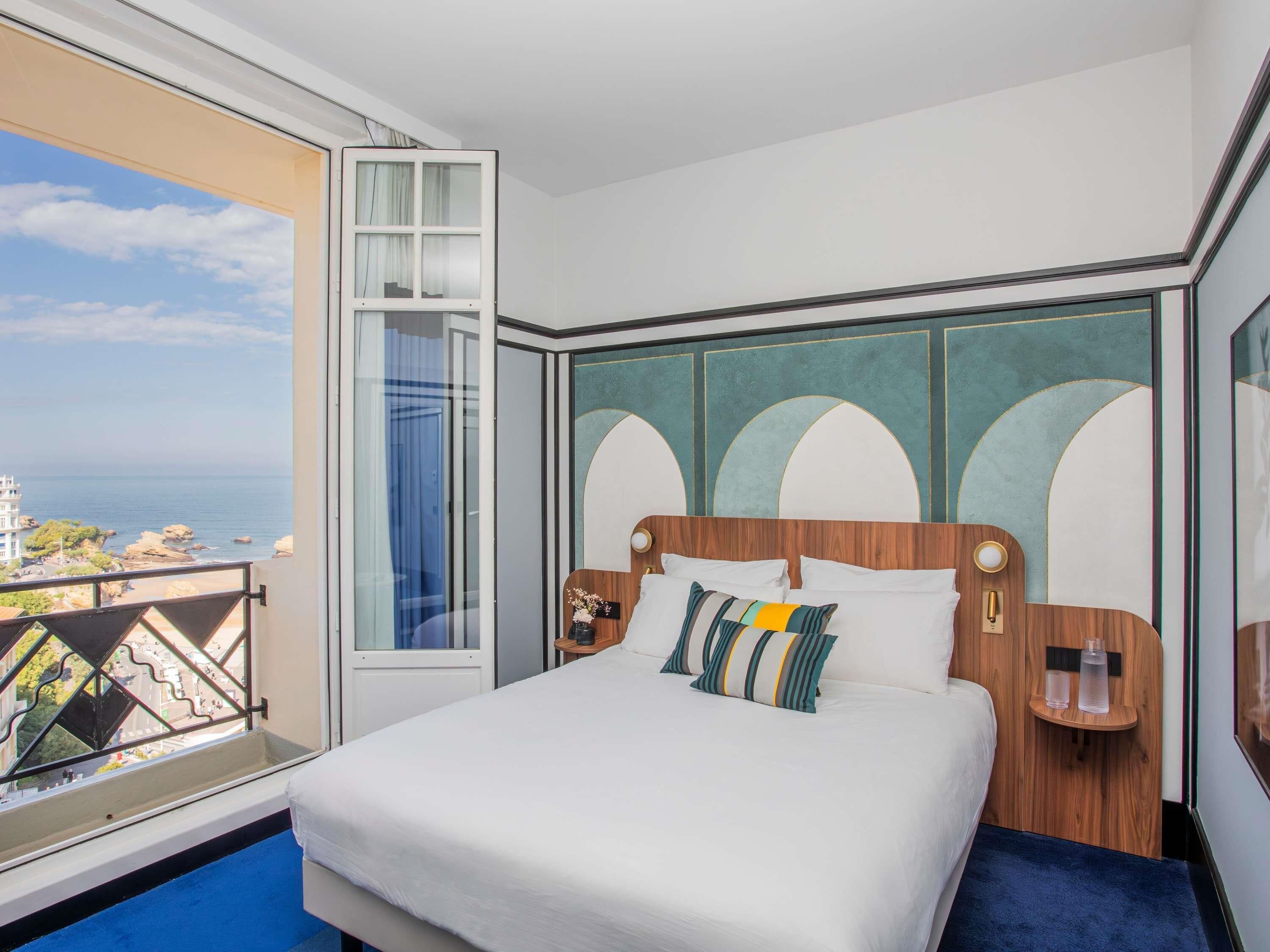 A hotel room with a large bed, a view of the sea, and a balcony with a railing.