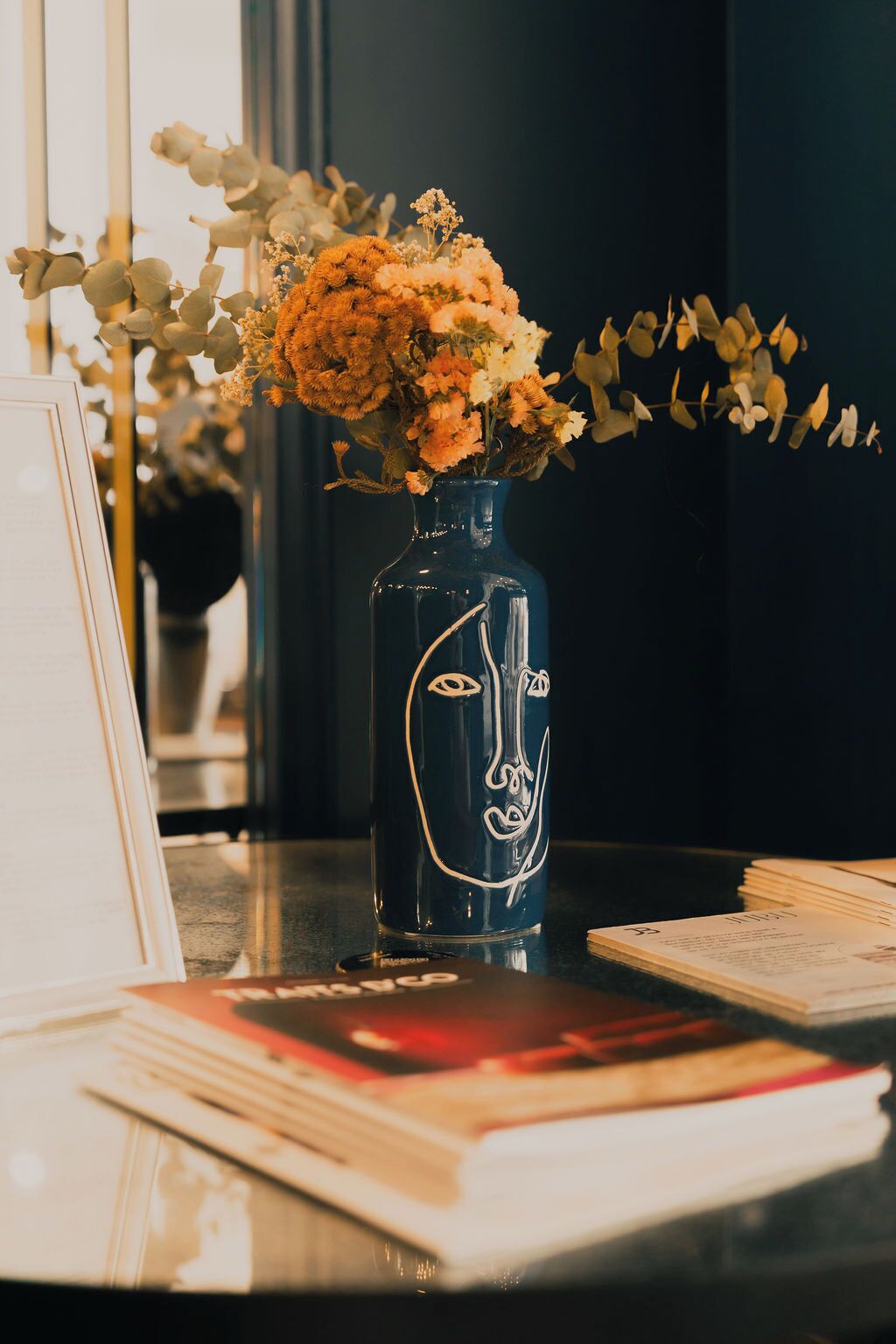 Eine Vase mit Gesichtsdekor auf einem Tisch mit Büchern und einem gerahmten Bild.