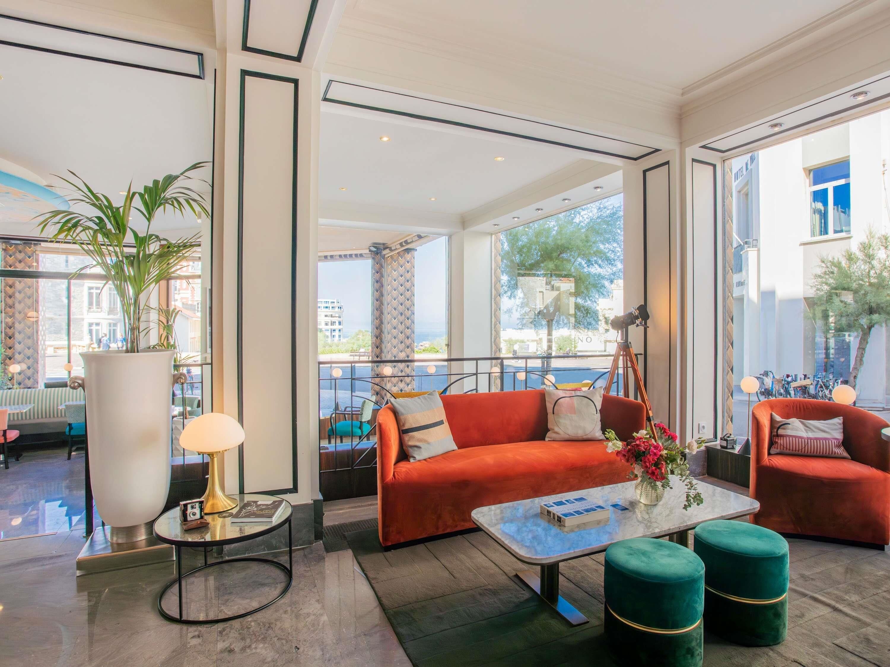 A modern hotel lobby with a red sofa, green ottomans, a glass coffee table, and a large potted plant.