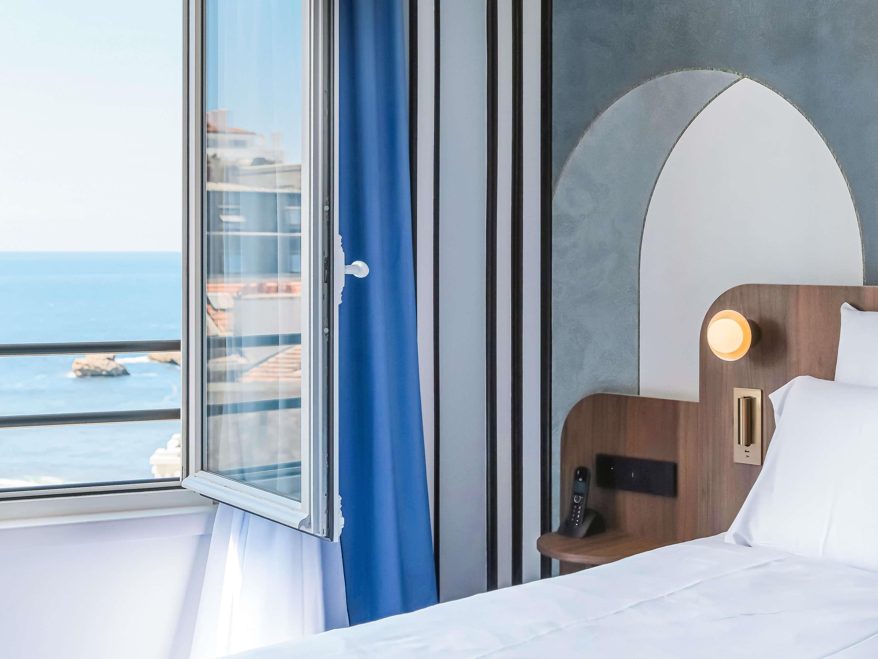 A hotel room with a bed, a bedside table with a phone, a window with curtains, and a view of the sea.