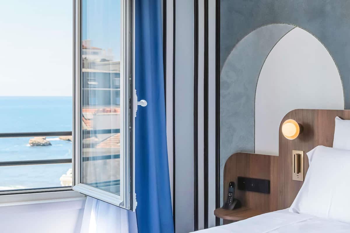 A hotel room with a bed, a bedside table with a phone, a window with curtains, and a view of the sea.