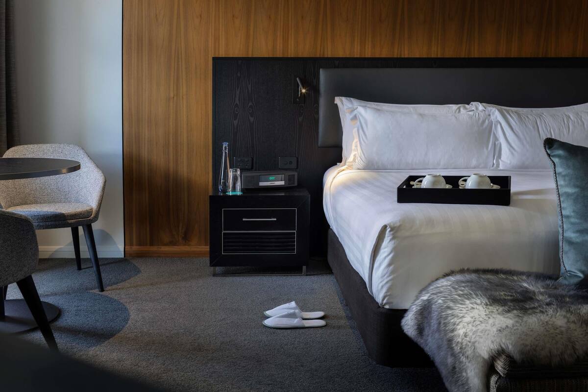 A modern hotel room with a bed, bedside table, chair, and a small table with a tray.
