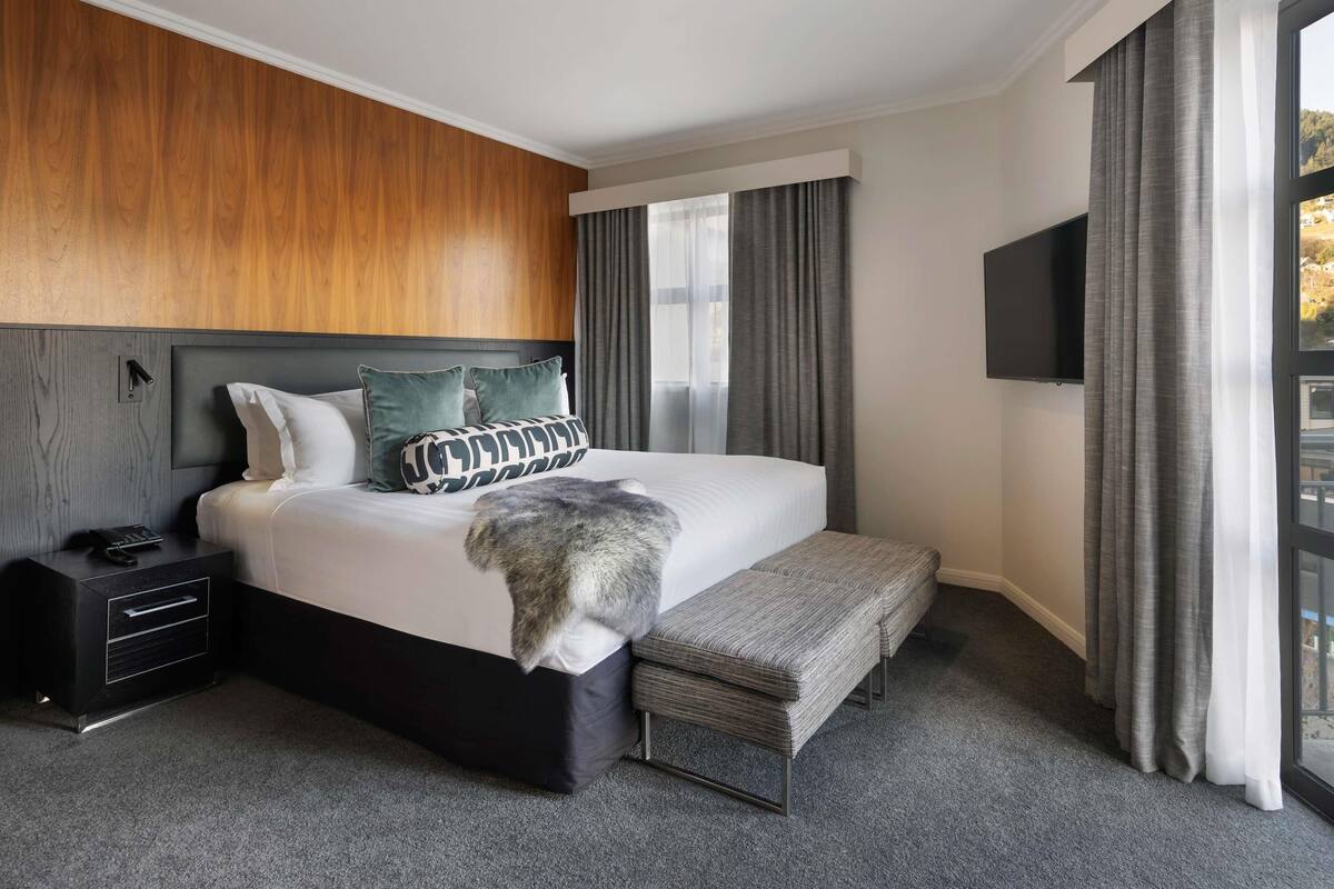 A modern hotel room with a large bed, a bedside table, a bench, a TV, and a window with curtains.