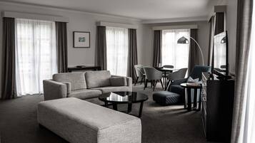 A modern hotel room with a sectional sofa, a round coffee table, a desk with a chair, and a television.
