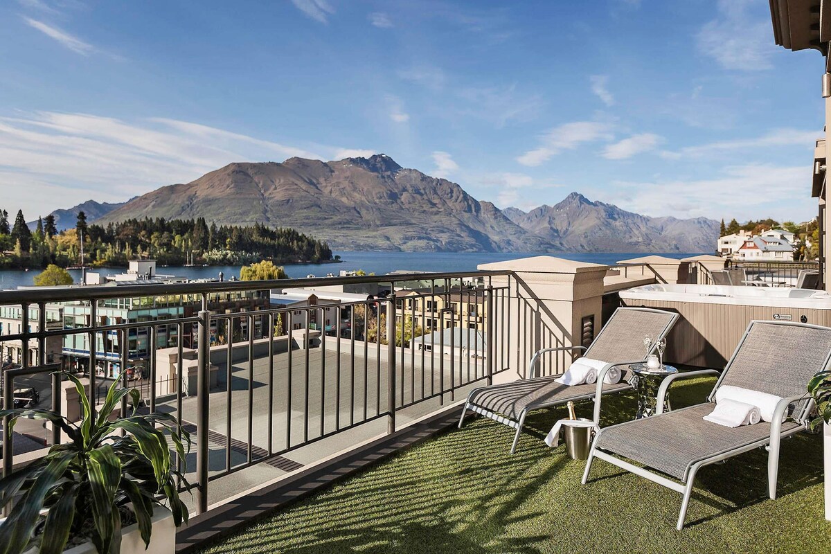 A balcony with a view of mountains and a lake, lounge chairs, and a railing.